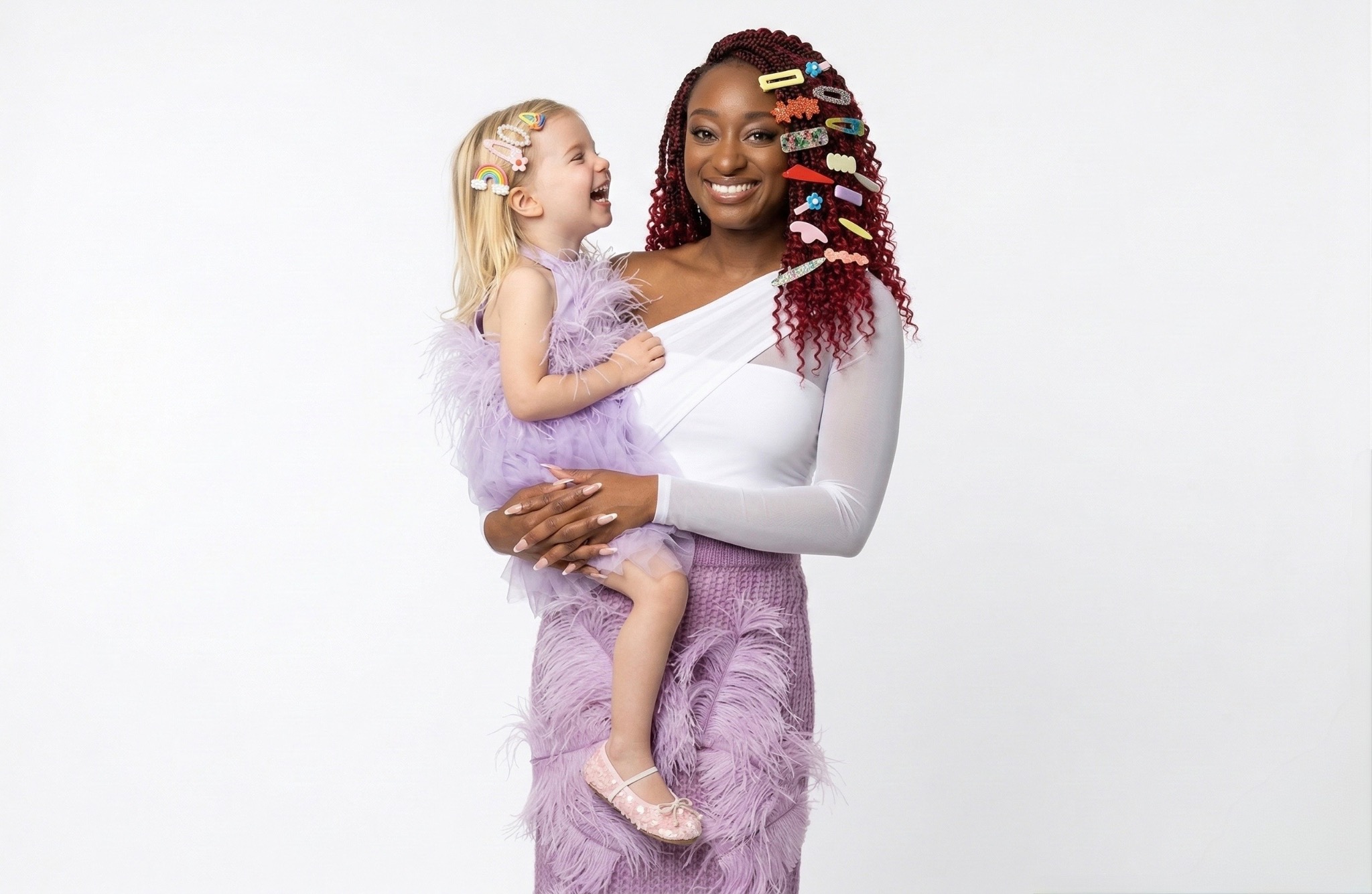 Shiloh in a white and purple skirt carrying a smiling little girl while wearing colorful hair clips